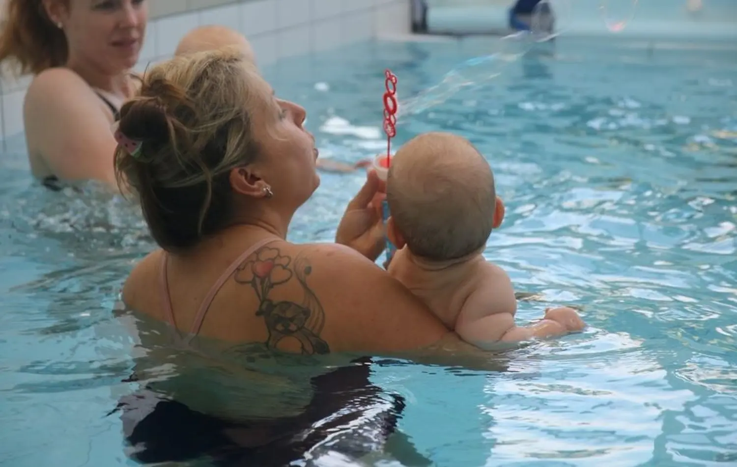 Swimmerfirst, Eine Frau in einem Pool hält ein Baby und bläst Seifenblasen